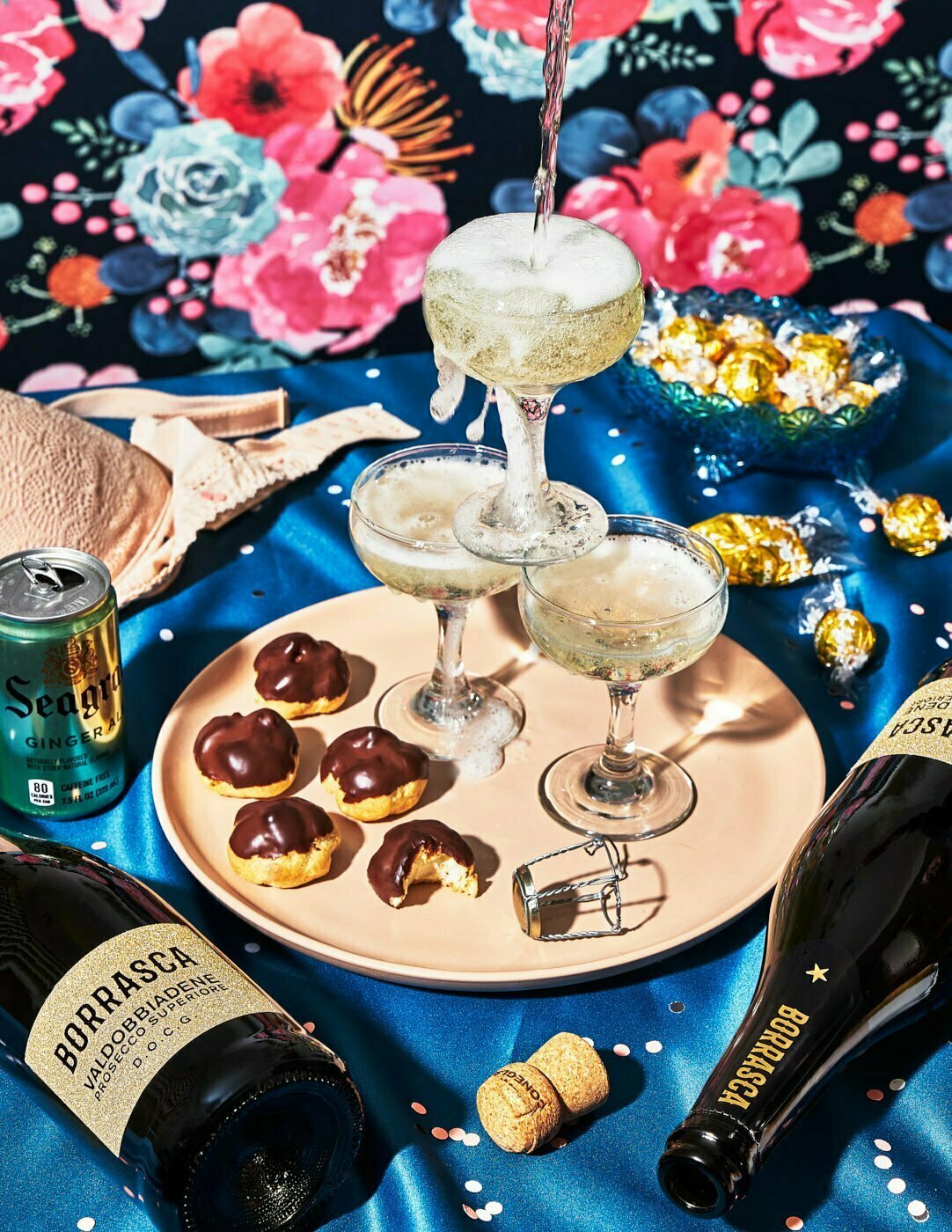 Open bottles of Borrasca Prosecco on a blue silk table surrounded bu half eaten snacks, confetti, and a stack of coupe champagne glasses with prosecco being pouring into the top glass.