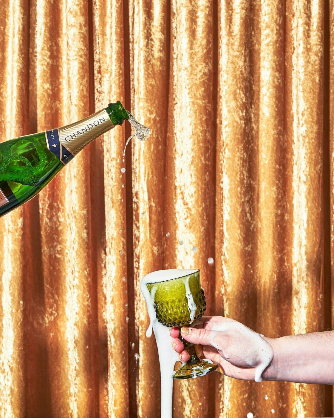 A bottle of Chandon champagne being poured into a green goblet that is over flowing with champagne.