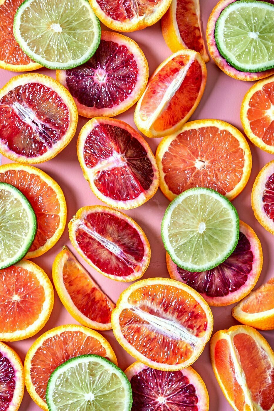 Sliced and quartered oranges, limes, and blood oranges sit on top of a pink background.