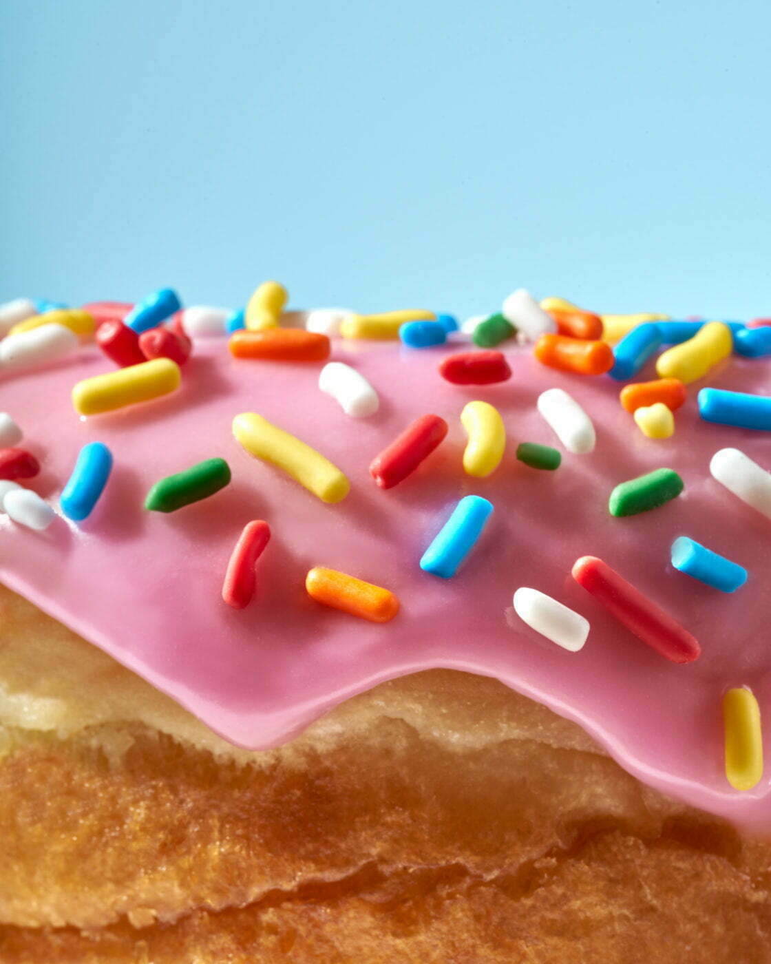 Close-up of a pink raised donut covered in rainbow sprinkles against a sky blue background.