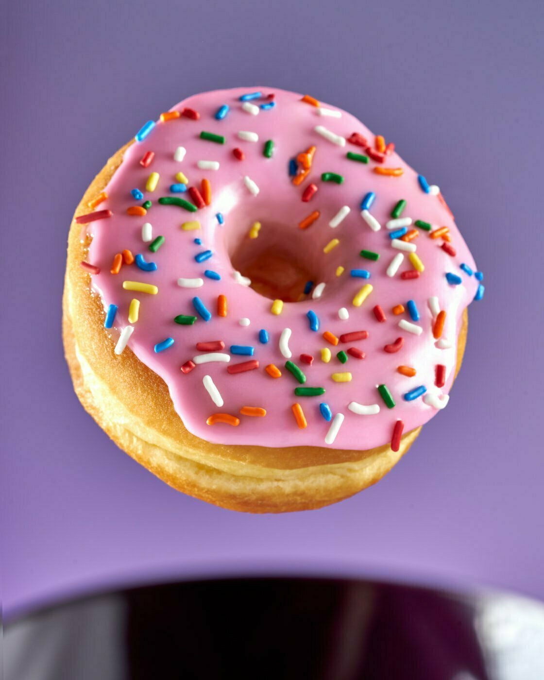 A pink raised donut with sprinkles floating above a purple mug against a purple background.