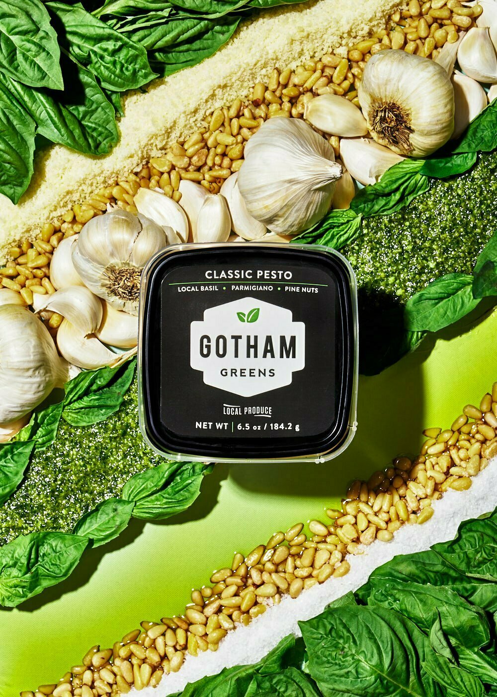 Gotham Greens Classic Pesto sits above a background of basil, parmesan, pine nuts, whole and garlic cloves, olive oil, and salt. Each ingredient is placed in a neat diagonal line behind the pesto.