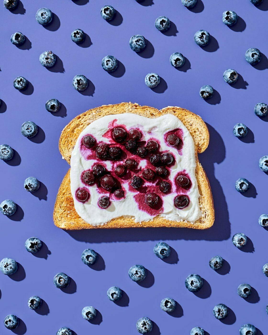 A single slice of whole wheat bread topped with greek yogurt and frozen blueberries on a purple background background with a pattern of fresh blueberries.
