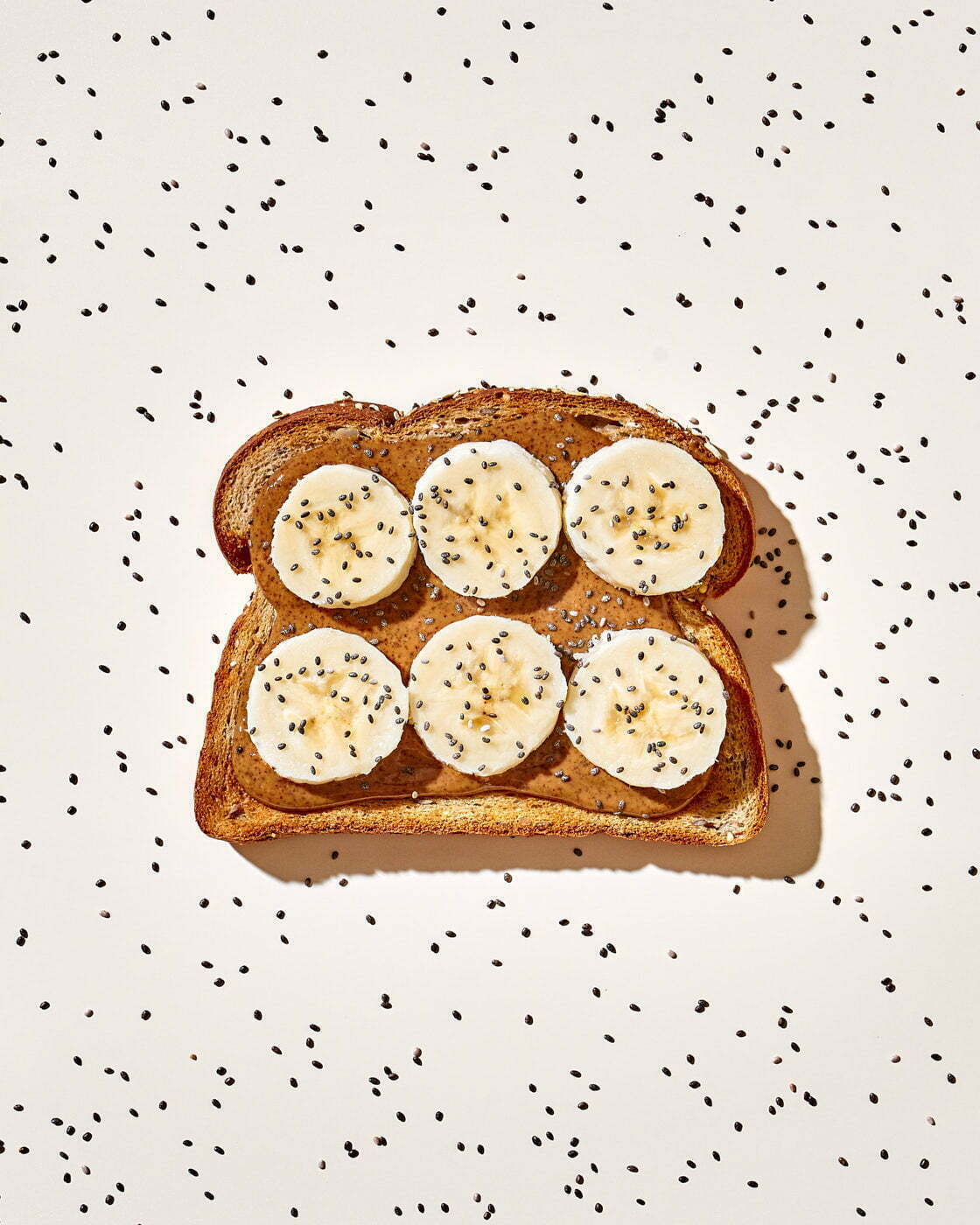 A single slice of whole wheat bread topped with almond butter, banana slices, and chia seeds on an off white background covered in chia seeds..