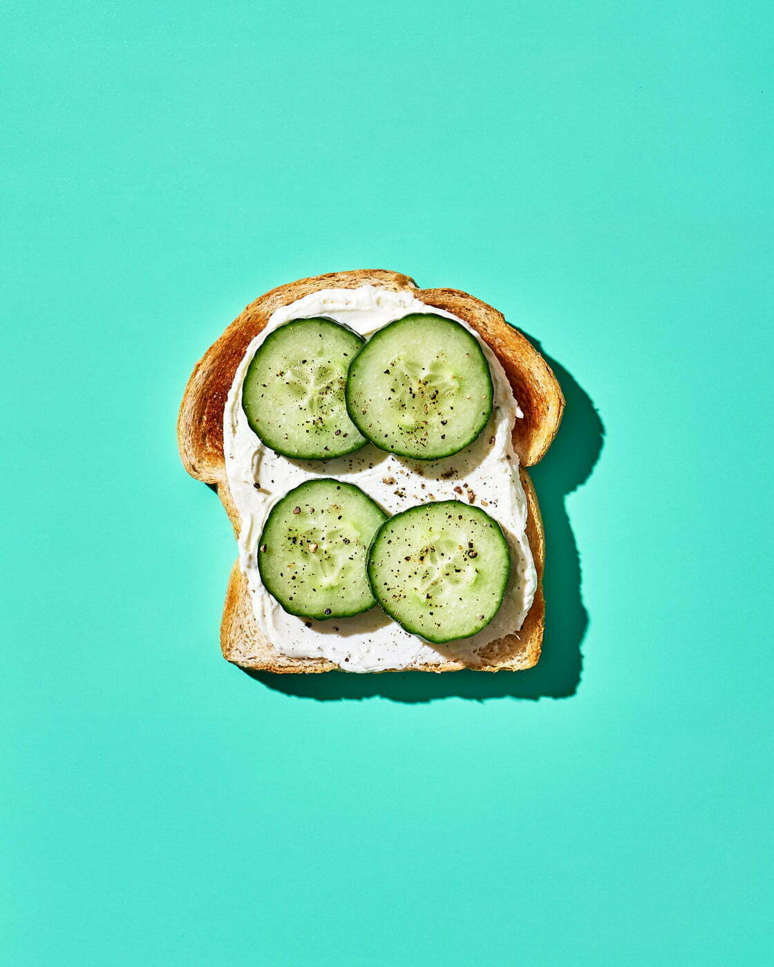 A single slice of sourdough bread topped with cream cheese, cucumbers, and freshly ground black pepper on a teal background.