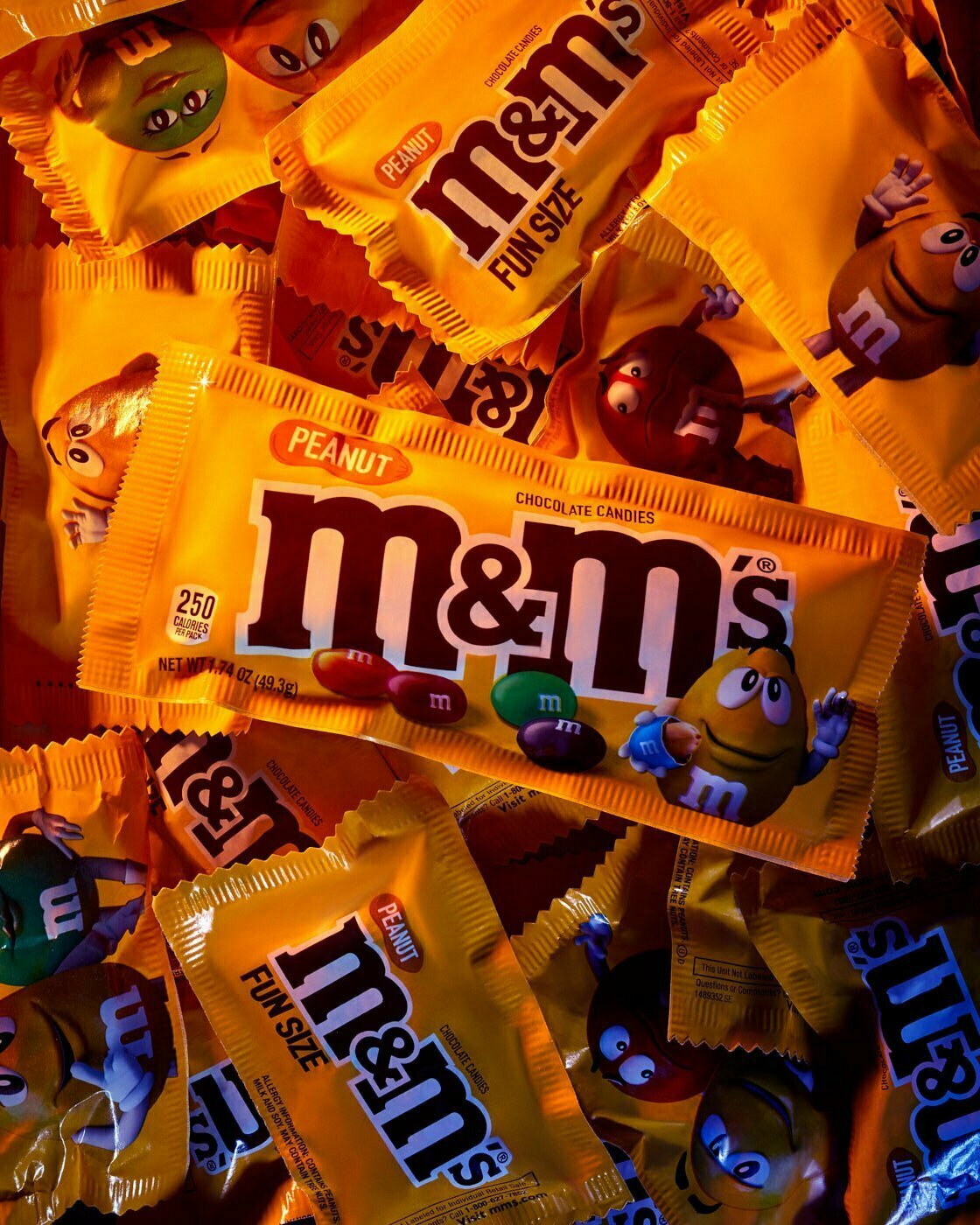A full size Peanut M&M candy still in wrapper sitting on top of mini Peanut M&M candy also still in their wrappers.