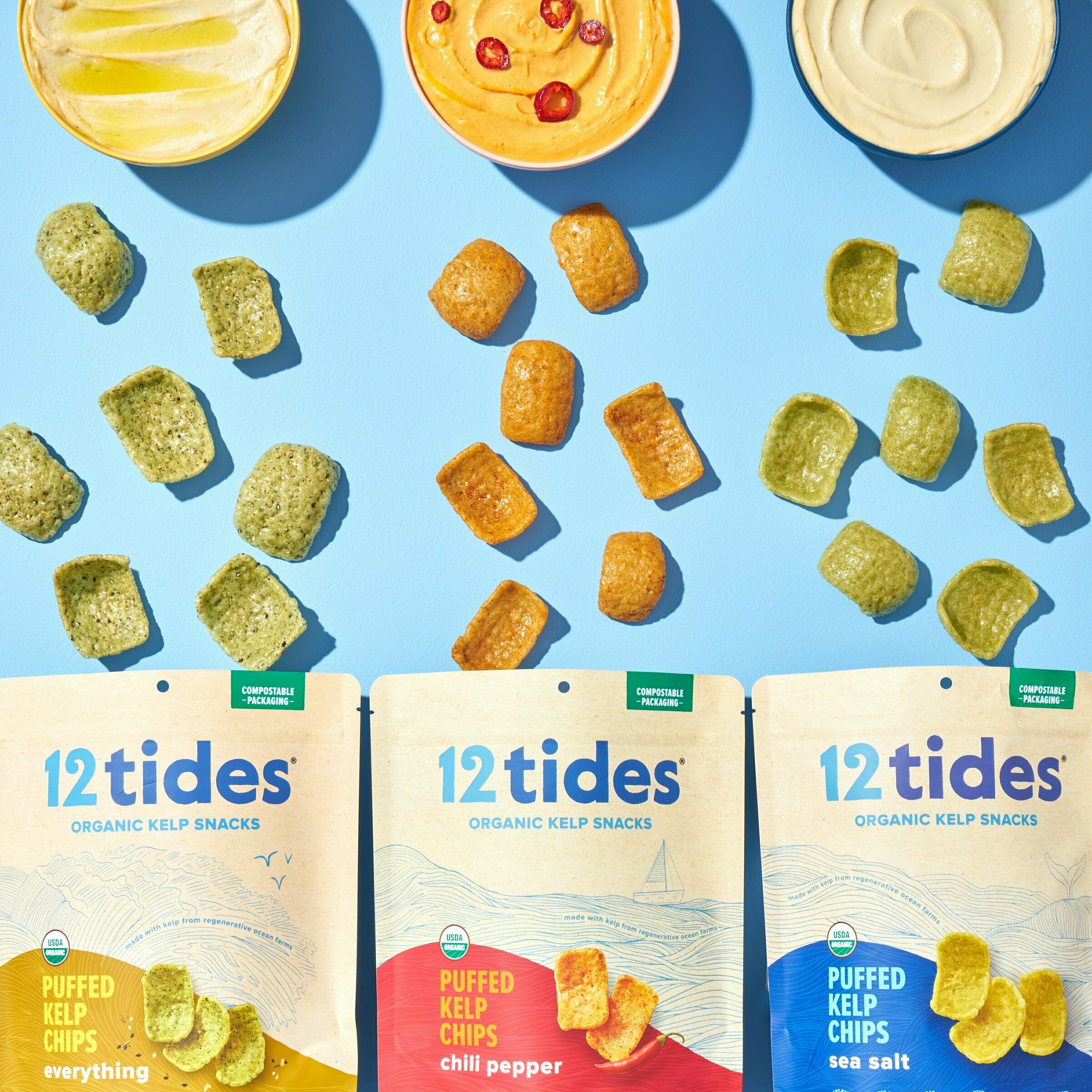 All flavors of 12 Tides Kelp Chips spilling out from each bag towards bowls of hummus